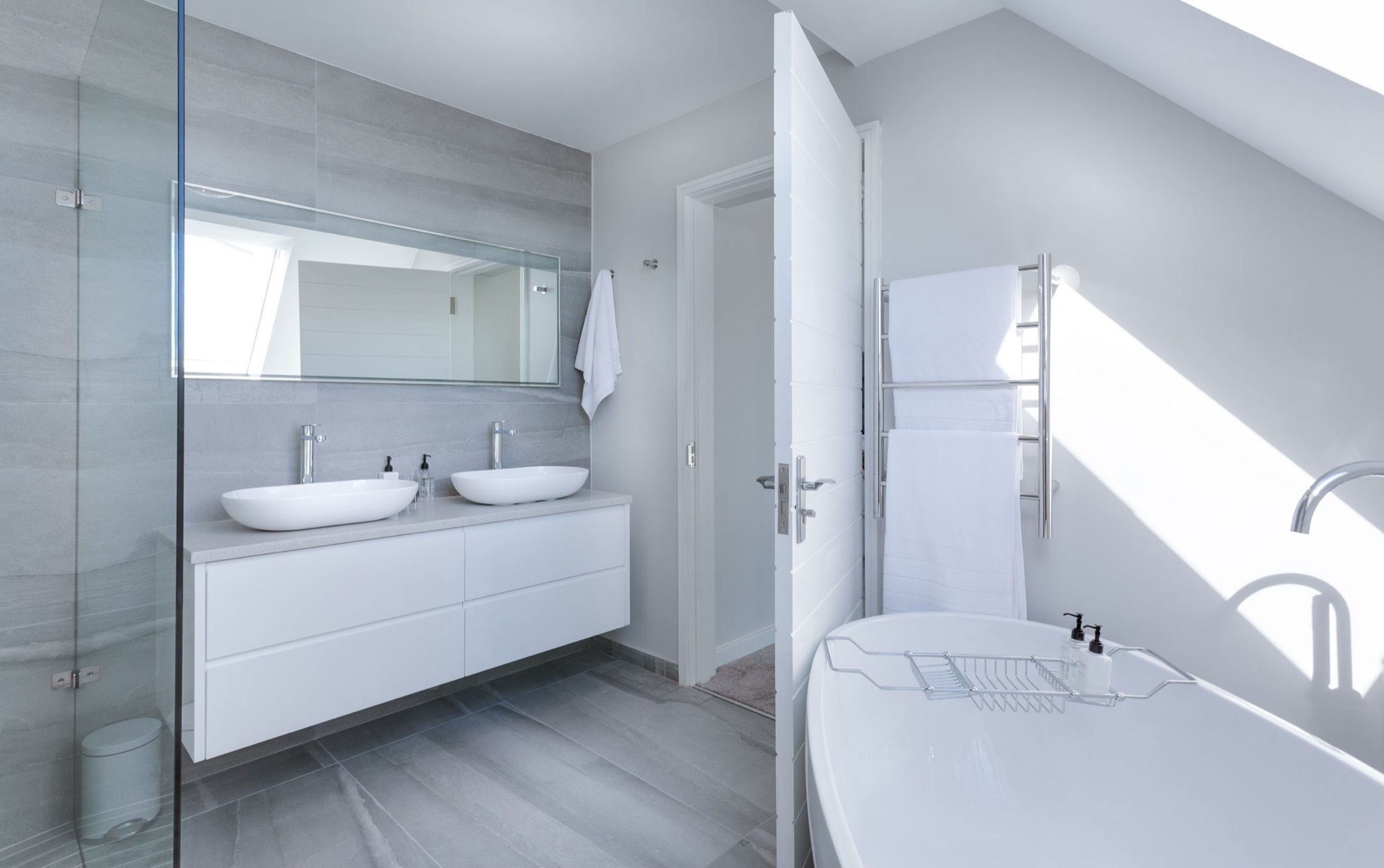 A light bathroom under a sloping ceiling with twin countertop basins on a wall-hung vanity, a heated towel rail and large stone-effect floor tiles
