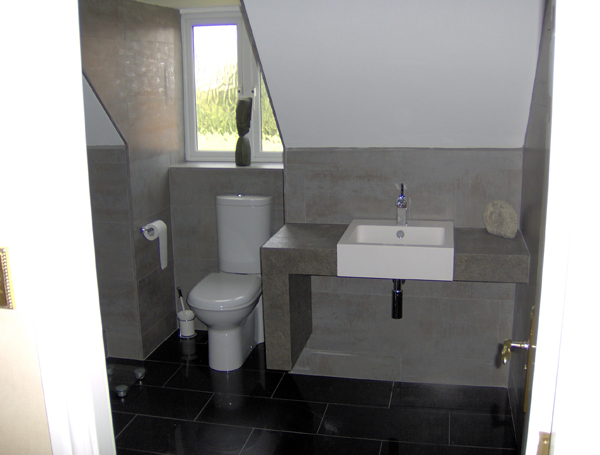 A downstairs cloakroom with a sloping ceiling, concrete-effect tiled walls, a countertop basin on a tiled shelf and a dark slate-effect floor