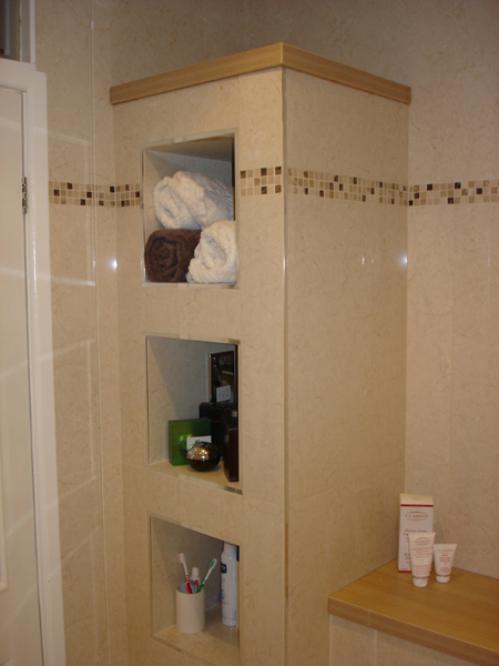 A tiled column in a bathroom with three built-in recessed niches holding towels and toiletries, with a mosaic tile border