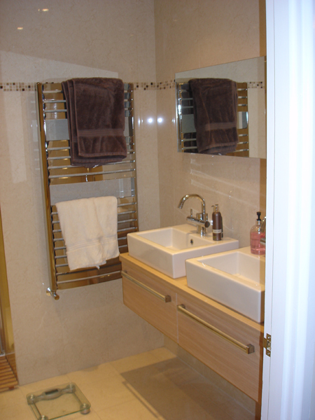 Twin white countertop basins on a wall-hung oak vanity unit beside a chrome heated towel rail hung with towels